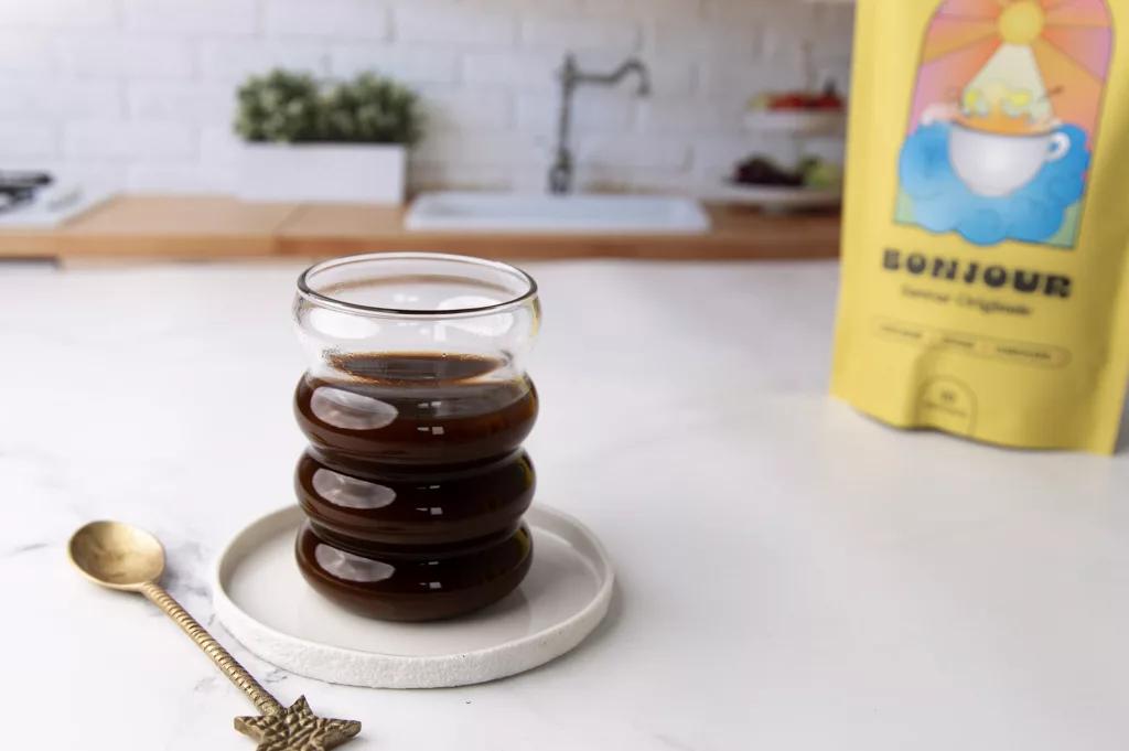 Tasse de boisson Bonour Drink dans un verre ondulé avec une cuillère dirée à sa gauche. En arrière plan un sachet jaune de la boisson Bonjour Drink