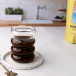 Tasse de boisson Bonour Drink dans un verre ondulé avec une cuillère dirée à sa gauche. En arrière plan un sachet jaune de la boisson Bonjour Drink