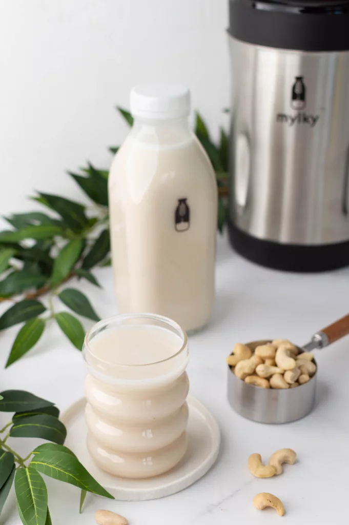 Un verre de lait de cajou en premier plan, des noix de cajou dans un récipient et La machine à lait végétal Mylky en arrière plan.