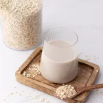 Que faire avec du lait d'avoine sainement Que faire avec du lait d'avoine. Photo d'un verre de lait d'avoine sur un petit plateau en bois, où une cuillère en bois de flocon d'avoine est posée coté. En arrière plan un bocal de flocon d'avoine