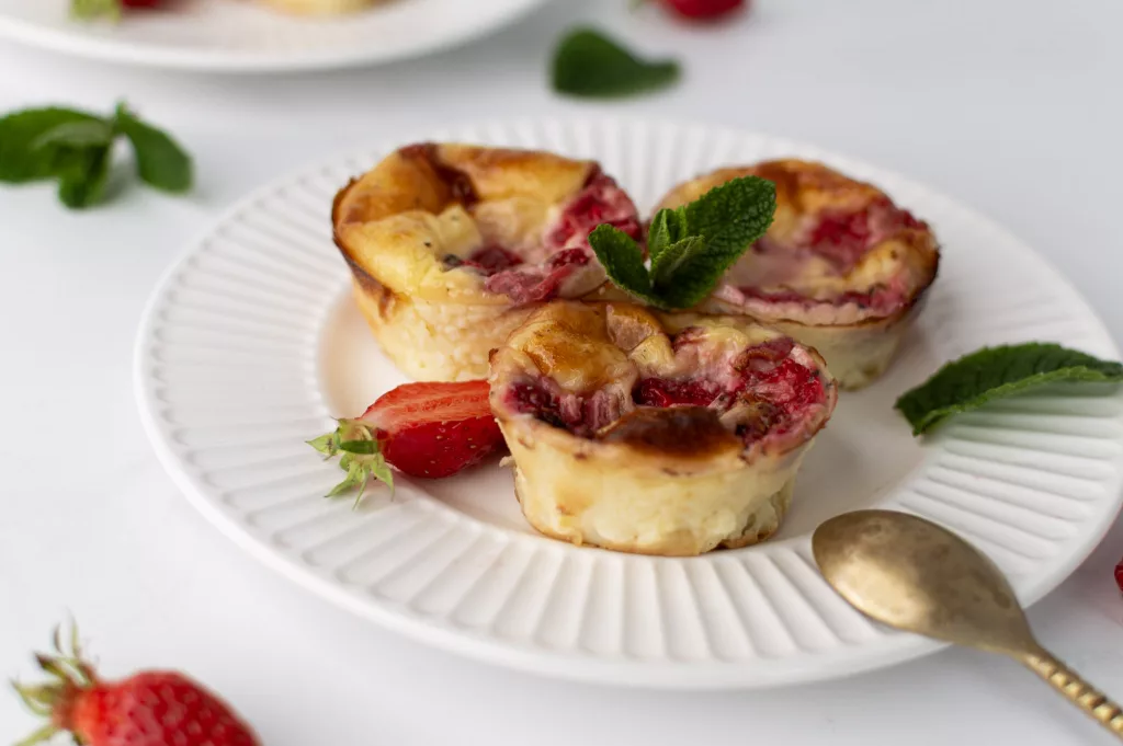3 petits flans aux fraises sur une assiette blznc. Le dos d'une petite cuillère dorée repose sur l'assiette. 1/2 fraise est posé sur l'assiette