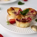 3 petits flans aux fraises sur une assiette blznc. Le dos d'une petite cuillère dorée repose sur l'assiette. 1/2 fraise est posé sur l'assiette