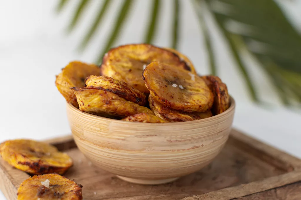 Banane plantain au four en rondelles dans une coupelle en bois avec une feuille de palmier en arrière plan