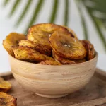 Banane plantain au four en rondelles dans une coupelle en bois avec une feuille de palmier en arrière plan