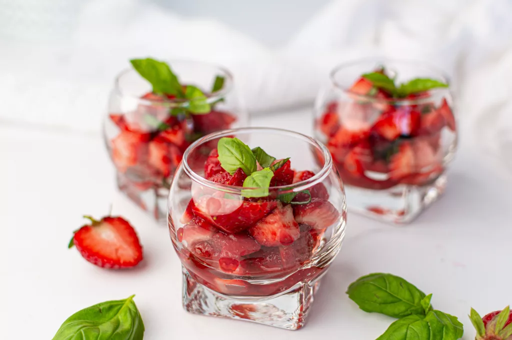 3 verrines remplies de salades de fraises et basilic sur fond blanc. Quelques fraises et feuilles de basilic tout autour