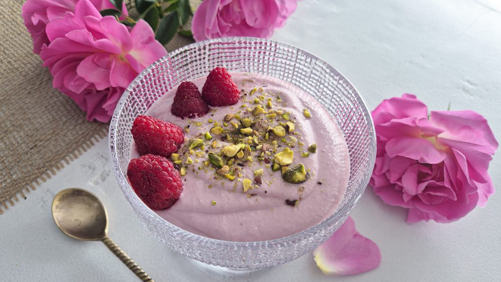 coupe de creme a la framboise et a la rose avec des framboises et des pistaches concassées sur le dessus. A l'arrière des roses roses et une cuillère couleur dorée